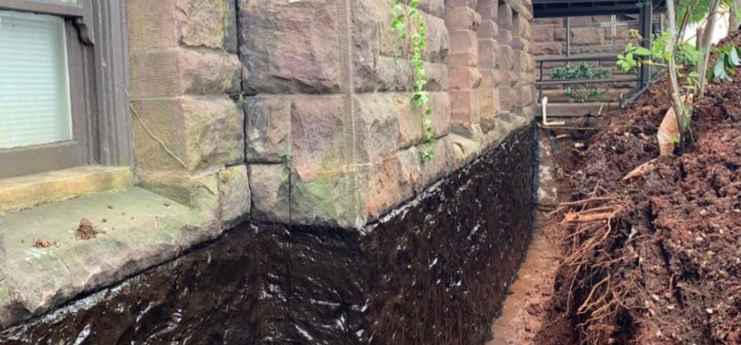Exterior foundation wall with fresh waterproofing sealant applied, showing an excavated trench around the home to prevent water intrusion and foundation damage.